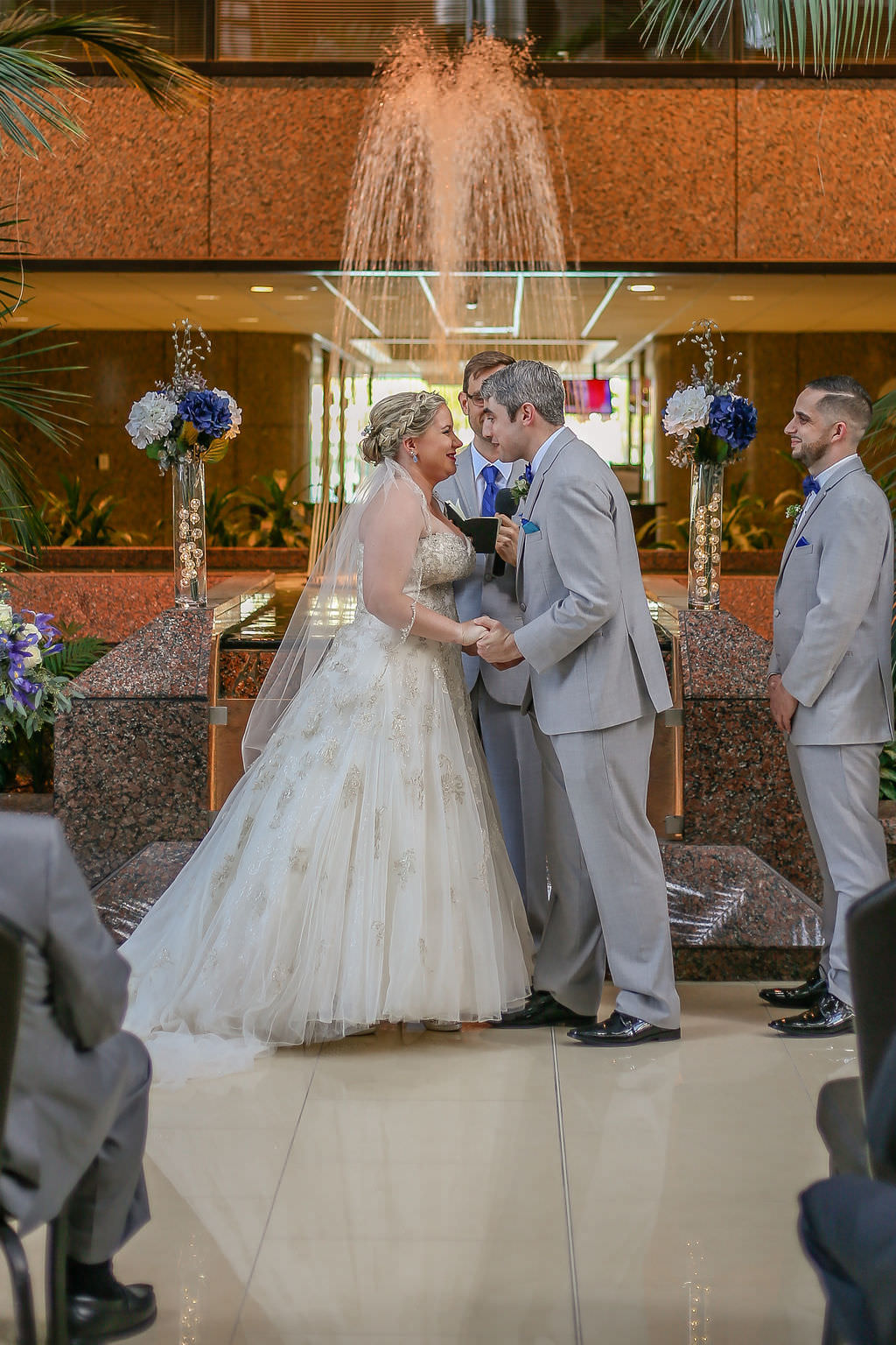 Elegant Indoor Florida Wedding Ceremony, Florida Bride and Groom at Altar, Bride Wearing Strapless A-Line Ivory Wedding Dress with Rhinestone Embellishments and Tulle Bottom , Groom in Light Gray Tuxedo, Waterfall Fountain in Ceremony Backdrop, White and Purple Wedding Ceremony Flowers | Tampa Bay Wedding Venue Tampa Centre Club