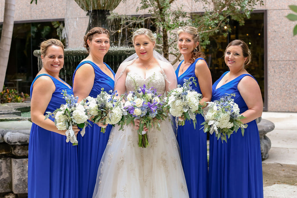 Florida Bride and Bridesmaids in Wedding Portrait, Bride wearing Strapless Sweetheart Neckline A-line Ivory Wedding Dress with Rhinestone Embellishments and Tulle Bottom, Ivory, Purple and Green Wedding Bouquet, Bridesmaids in David’s Bridal Sleeveless V-Neckline Long Blue Bridesmaid Dresses, In Front of Fountain | Tampa Bay Wedding Venue Tampa Centre Club