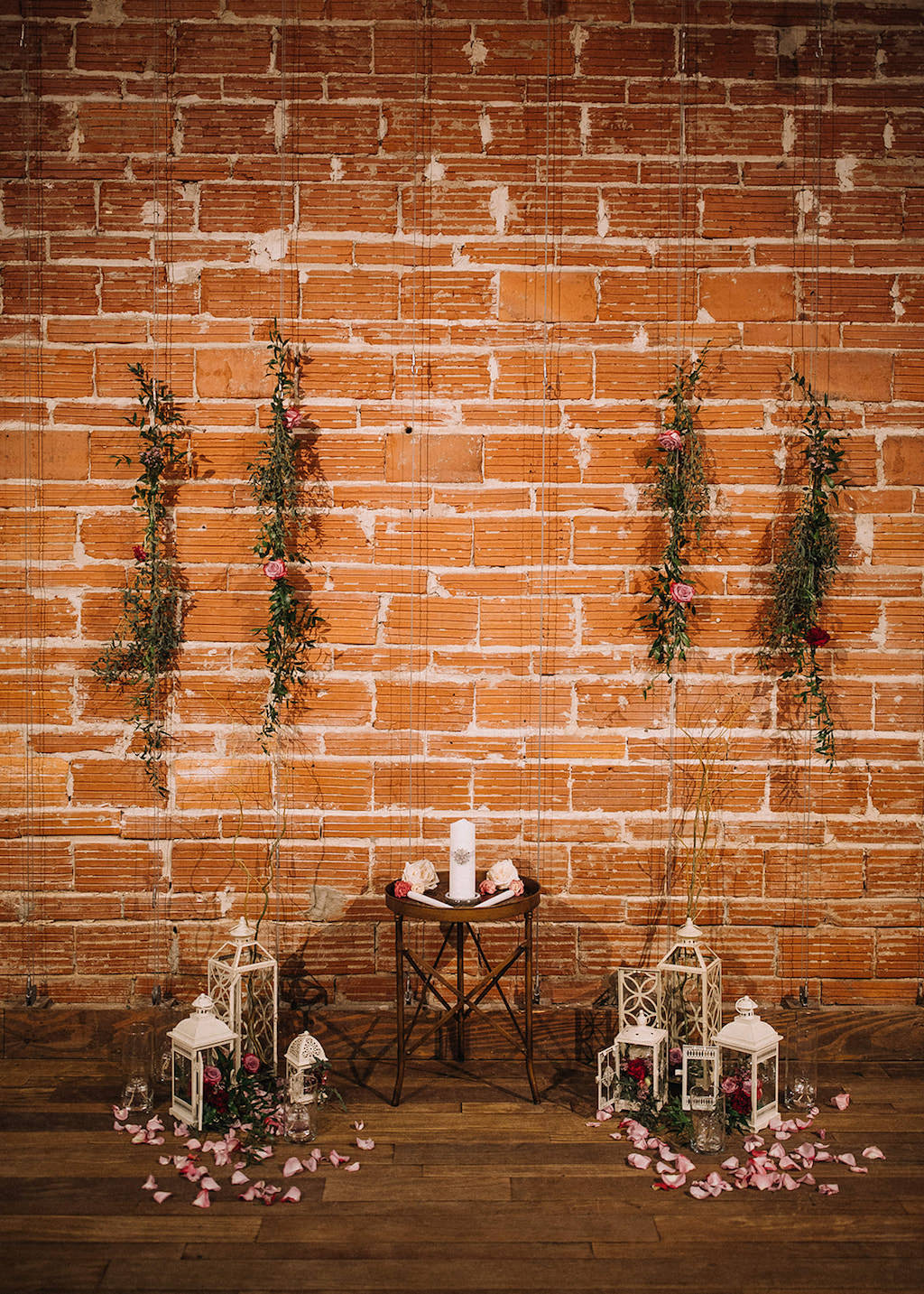 Romantic Boho Ceremony Backdrop, White Unity Candle, Vintage White Lanterns with Red Roses and Glass Liquid Candles, Hanging Greenery and Roses in front of Red Exposed Brick Wall | Downtown St. Pete Wedding Venue NOVA 535
