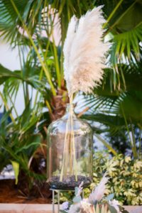 White Pampas Grass In Oversized Glass Water Jug with Black Pebbles On Top Of Natural Wooden Decor at Wedding Ceremony | St. Pete Beach Resort and Hotel Wedding Venue The Don Cesar | Tampa Bay Wedding Photographer Andi Diamond Photography | Tampa Bay Wedding Planner UNIQUE Weddings + Events