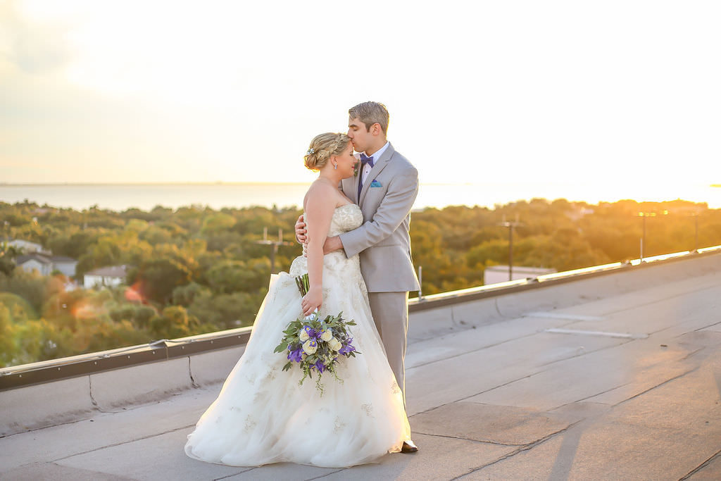 Florida Bride and Groom Rooftop Wedding Portrait at Sunset with Water Views, Bride wearing Strapless A-line Ivory Wedding Dress with Rhinestone Embellishments and Tulle Bottom, Bridal Updo with Braid, Ivory Purple and Green Wedding Bouquet, Groom in Light Gray Tuxedo with Blue BowTie | Tampa Bay Wedding Venue Tampa Centre Club