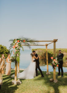 Florida Bride and Groom Intimate Wedding Photo Under Bamboo in Waterfront Ceremony, Bride in White Spaghetti Strap Beaded Karen Willis Holmes Wedding Dress with Veil, Tropical Wedding Decor with Colorful Flowers, Pink Ginger, King Proteas, Orchids, with Palm Leaf Greenery and Pampas Grass | Tampa Bay Beachfront Wedding Venue Longboat Key Club