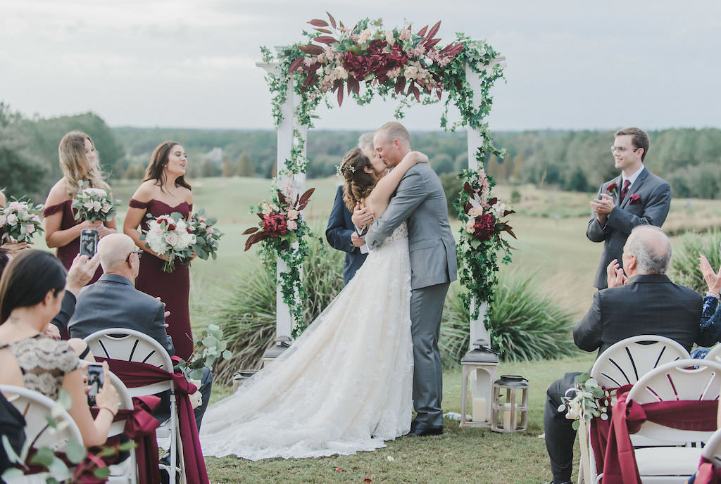 Florida Bride and Groom Kiss During Rustic Chic Outdoor Wedding Ceremony, Under White Arch with Burgundy, Blush Pink and White Floral Arrangements with Greenery | Brooksville Golf Course Southern Hills Plantation | Tampa Bay Bridal Shop Nikki’s Glitz and Glam Boutique