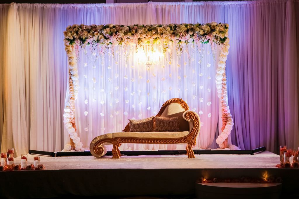 Traditional Indian Wedding Reception Decor, White Linen Backdrop with Purple Uplighting, Ivory danders Gold Couch, Extravagant White, Blush Pink Floral Arrangements on Arch