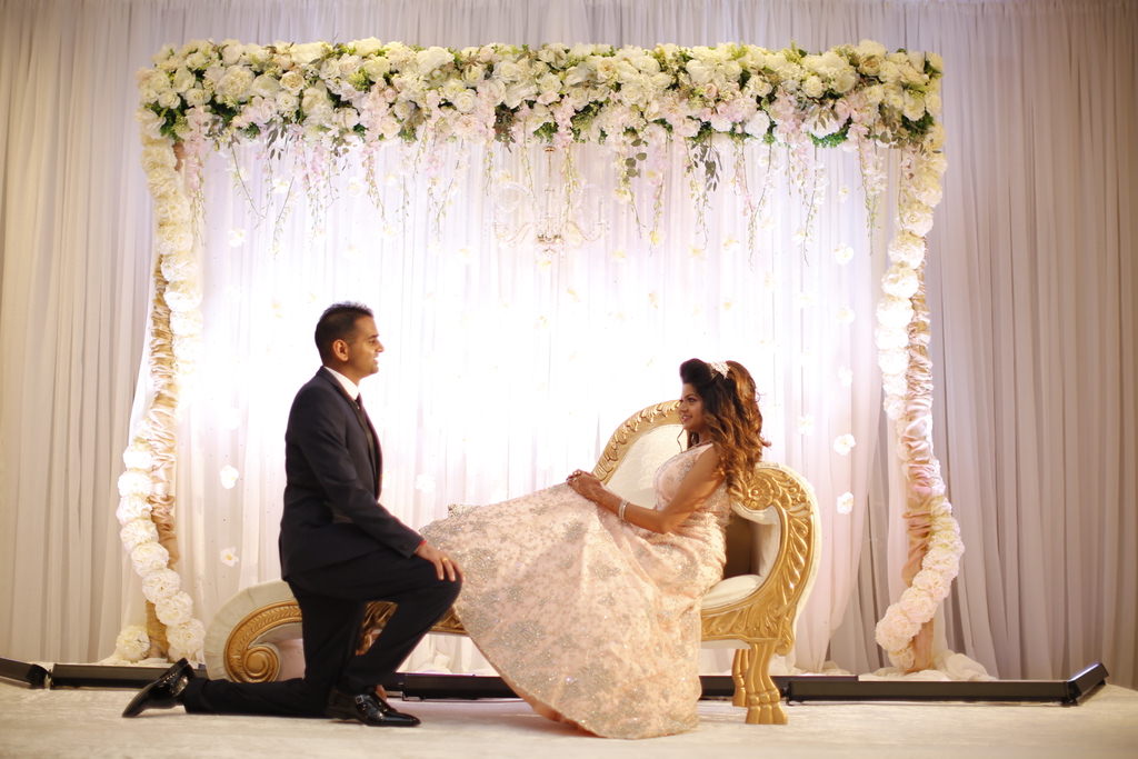 Tampa Elegant Traditional Indian Bride and Groom, White Linens Backdrop, Extravagant White, Ivory and Blush Pink Floral Arch, Bride Lounging on White and Gold Couch in White Dress, Groom Kneeling in Black Suit
