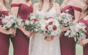 Florida Bridal Party Portrait, Bridesmaids in Long Burgundy Bill Levkoff Dresses with Off The Shoulder Sleeves, Carrying Red Carnations, Blush Pink Roses, and White Floral Bridal Bouquets with Silver Dollar Eucalyptus Greenery | Tampa Bay Bridal Shop Nikki’s Glitz and Glam Boutique