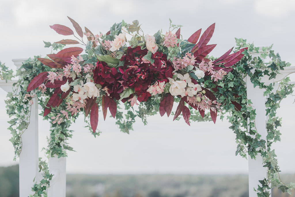 Romantic FSU Inspired Wedding Decor, Burgundy, Blush Pink, and White Floral Arrangement on Top of White Wedding Arch with Greenery and Garland