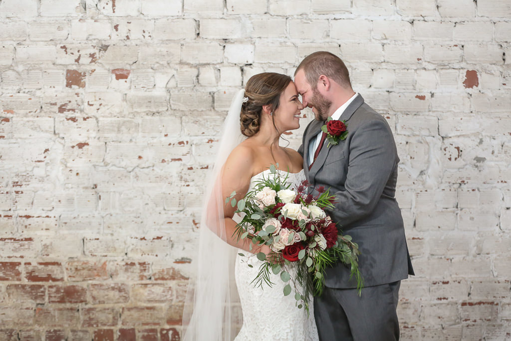 Tampa Bay Groom and Bride in Strapless Lace Fitted Wedding Dress and Cathedral Length Veil with Organic Red, Blush, Ivory and Greenery Floral Bouquet and White Brick Backdrop | Tampa Wedding Photographer Lifelong Photography Studios | Industrial Historic Wedding Venue Rialto Theatre | Tampa Wedding Hair and Makeup Artists Michele Renee the Studio