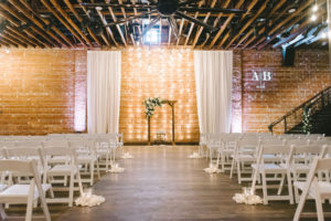 Modern, Rustic Elegant Indoor Ceremony, Exposed Red Brick Wall, White Draping and Hanging Lights, Wooden Ceremony Arch with White and Greenery Floral Arrangement, White Folding Chairs, Custom Monogram Gobo Projection Wedding Decor | Downtown St. Pete Venue NOVA 535 | Tampa Bay Photographer Kera Photography