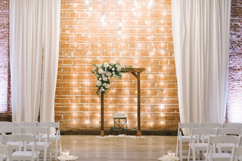 Modern, Rustic Romantic Indoor Ceremony, String Light Backdrop, Exposed Red Brick Wall, White Wedding Decor and Draping, Wooden Arch with White and Greenery Floral Arrangement | Downtown St. Pete Venue NOVA 535 | Tampa Bay Photographer Kera Photography