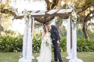 Palm Harbor Bride and Groom Kissing Wedding Portrait Under Wooden Ceremony Arch with White Draping and Ivory and Blue Flower Bouquets with White Flower Petals Blowing in the Wind | Tampa Bay Wedding Photographer Kristen Marie Photography | Innisbrook Golf & Spa Resort Wedding Venue