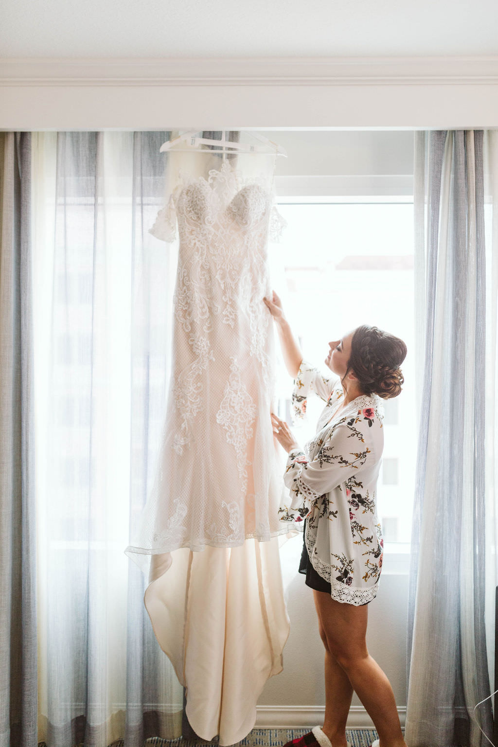 Florida Bride Getting Ready Wedding Portrait in White Floral Robe Looking at Lace Fitted Off the Shoulder Illusion Neckline Wedding Dress