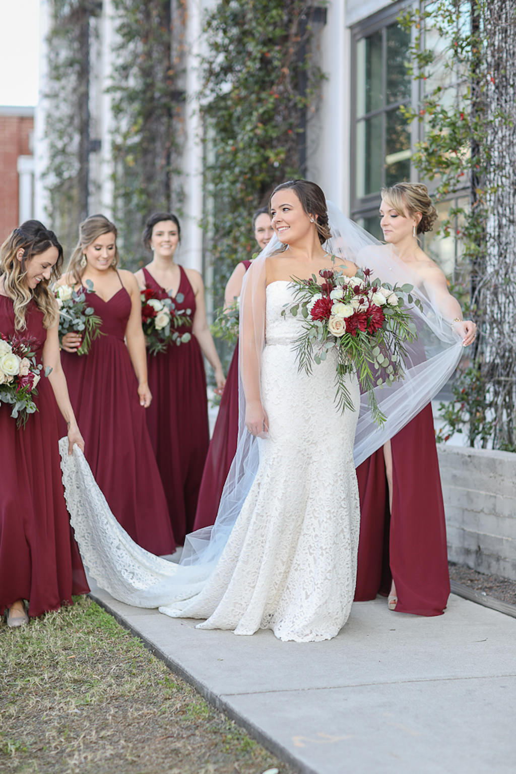 Bride in Strapless Lace and Fitted Wedding Dress with Rhinestone Belt and Cathedral Length Veil and Bridesmaids in Long Burgundy Wine Red Dresses Holding Garden Style Red, Ivory, and Greenery Floral Bouquets | Tampa Bay Wedding Photographer Lifelong Photography Studios | Tampa Bay Wedding Planner Breezin' Weddings | Tampa Bay Wedding Hair and Makeup Team Michele Renee the Studio