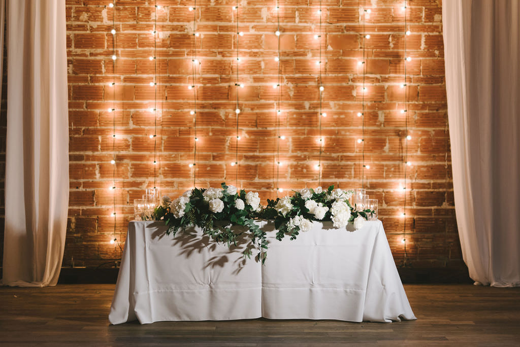 Elegant, Modern White, Gold and Greenery Wedding Reception Sweetheart Table Decor, Low Floral Centerpiece, String Light and Red Brick Backdrop | Downtown St. Pete Venue NOVA 535 | Tampa Bay Photographer Kera Photography