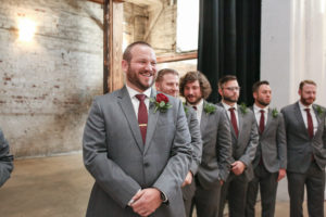 Groom Expression Watching Bride Walking Down Ceremony Aisle and Groomsmen in Grey Suits with Burgundy Ties and Red Rose Boutonniere | Tampa Wedding Photographer Lifelong Photography Studios