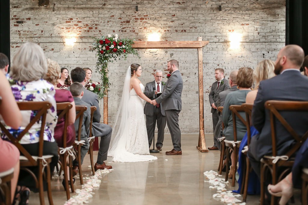 Tampa Bay Bride and Groom Exchanging Vows Under Wooden Arch with Red, Ivory and Greenery Floral Accent Wedding Ceremony Portrait | Tampa Bay Wedding Photographer Lifelong Photography Studios | Tampa Wedding Planner Breezin' Weddings | Historic Wedding Venue Rialto Theatre