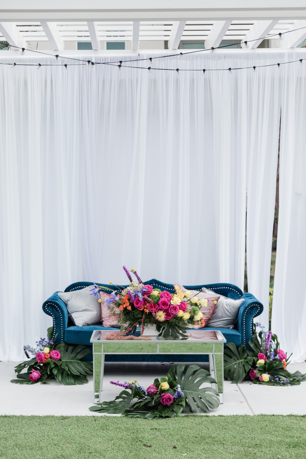 Modern, Tropical Garden Inspired Wedding Decor, Blue Velvet Lounge Seating and Couch, Dusty Rose Gold and Silver Accent Pillows, Mirrored Table, Whimsical Pink, Yellow, Orange, Purple Floral Centerpiece with Green Monstera Leaves, Against White Draping with Romantic Outdoor String Lighting | Tampa Bay Wedding Planner Special Moments Event Planning | Tampa Bay Wedding Florist Gabro Event Services