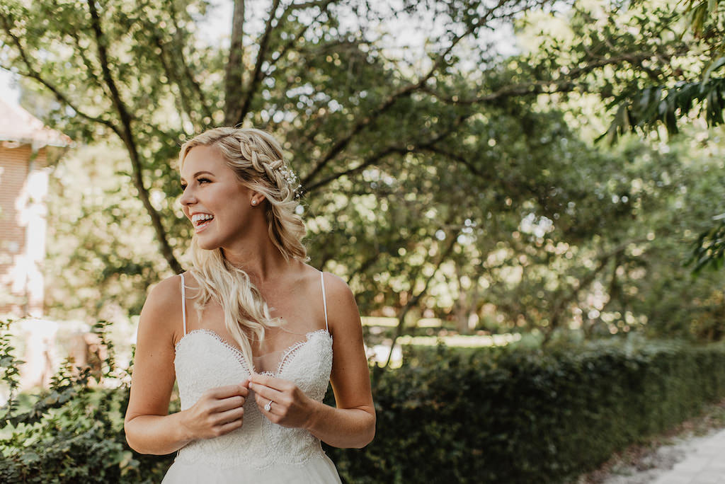 Fun, Happy Florida Bride Smiling Outdoor Garden Beauty Wedding Portrait, Spaghetti Strap Lace Bodice Sweetheart Neckline Wedding Dress and Braided Curled Half Up Do with Baby Breathes Floral Accessory | Tampa Bay Wedding Hair and Makeup Femme Akoi