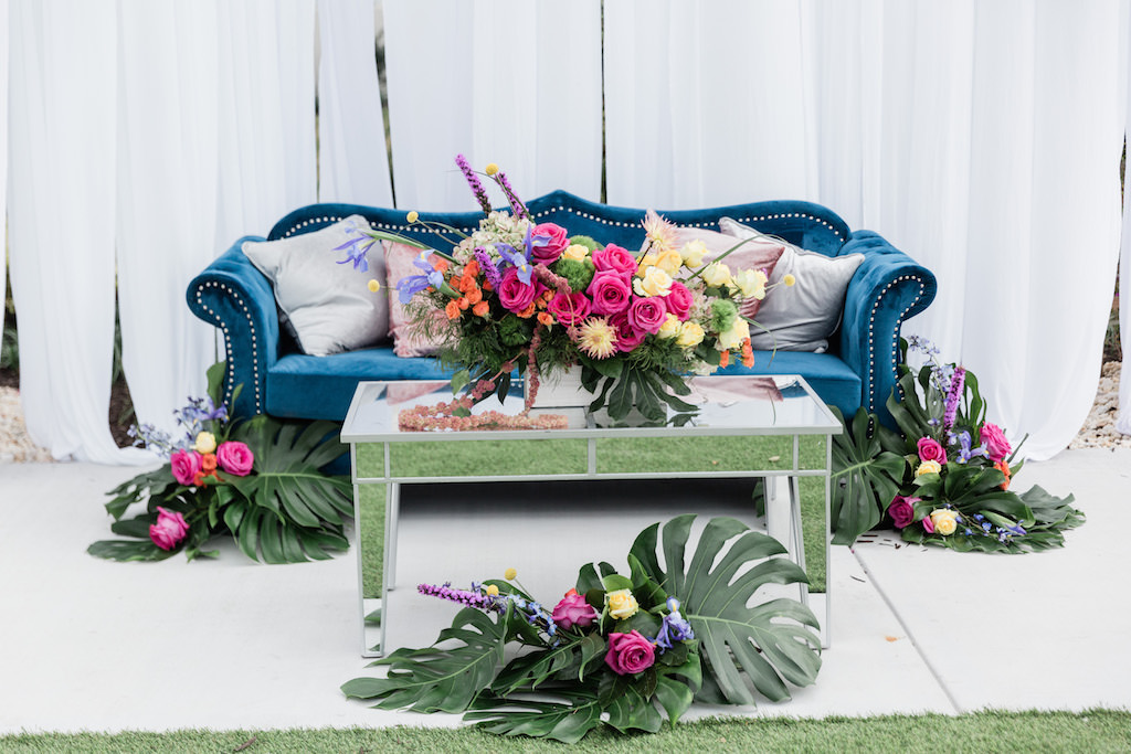 Modern, Tropical Garden Inspired Wedding Decor, Blue Velvet Lounge Seating and Couch, Dusty Rose Gold and Silver Accent Pillows, Mirrored Table, Whimsical Pink, Yellow, Orange, Purple Floral Centerpiece with Green Monstera Leaves, Against White Draping | Tampa Bay Wedding Planner Special Moments Event Planning | Tampa Bay Wedding Florist Gabro Event Services