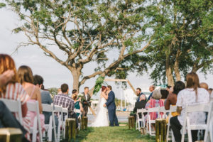 Florida Bride and Groom Exchanging First Kiss During Wedding Ceremony Under Modern Unique Wooden Geometric Ceremony Arch with Tropical Monstera Palm Leaves | Wedding Photographer Kera Photography | St. Petersburg Outdoor Waterfront Wedding Venue The Poynter Institute