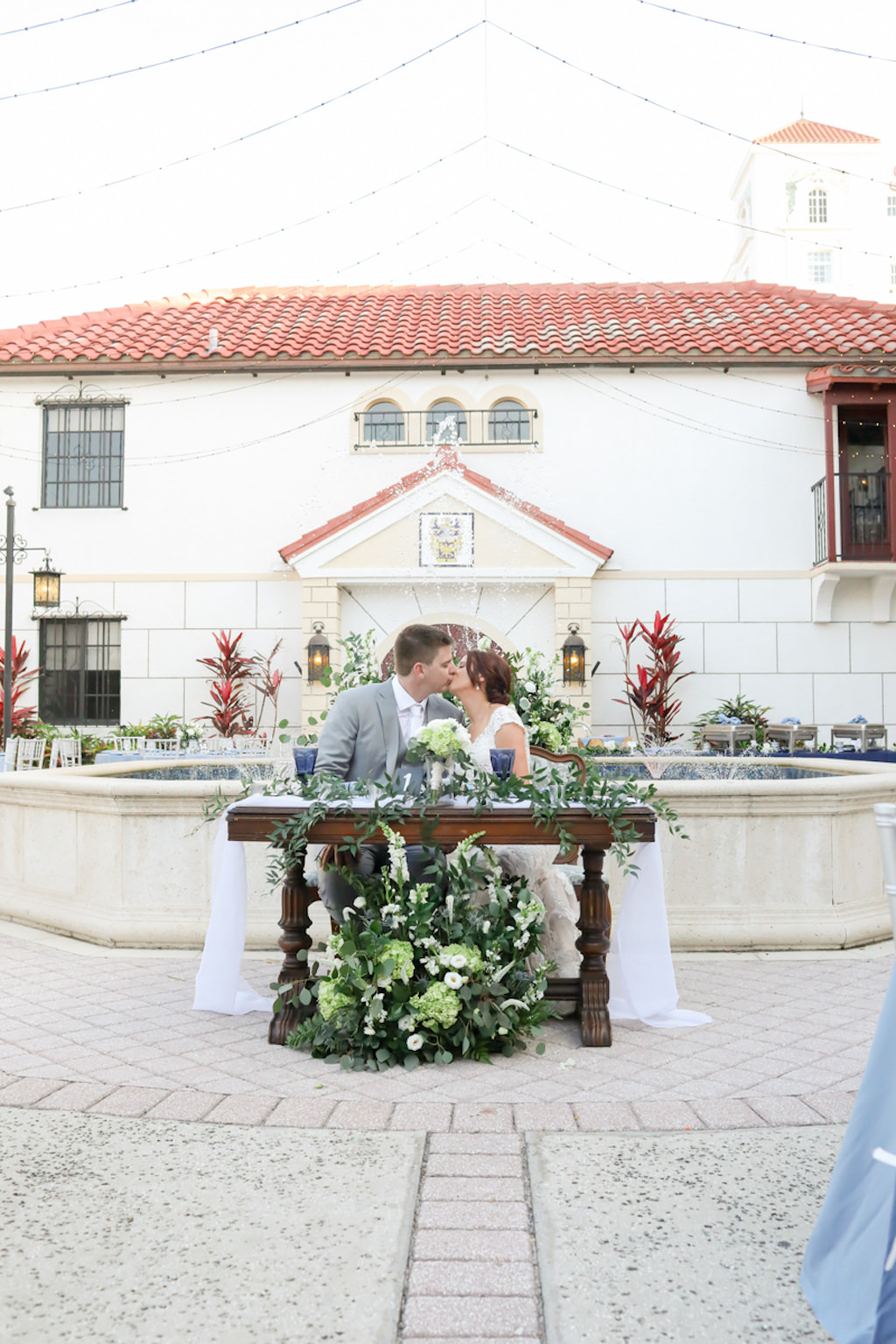 Tampa Bay Bride and Groom at Sweetheart Table Courtyard Wedding Reception Portrait, Wooden Table with Rustic Elegant Greenery, White, and Ivory Floral Arrangements | Wedding Photographer Lifelong Photography Studios | Wedding Planner Kelly Kennedy Weddings and Events | Wedding Venue Bishop Museum of Science and Nature