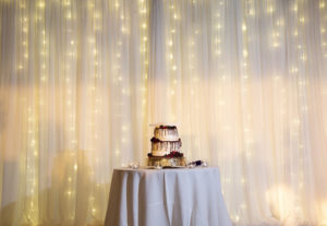 Elegant Two Tier White Wedding Cake, Chocolate Drip Frosting, Berries, White Lovebirds Cake Topper, White Draping with Romantic String Lighting Backdrop | Tampa Wedding Planner Laura Detwiler Events