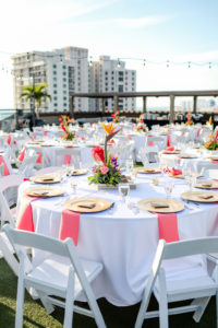Tropical, Florida Beach Inspired Wedding Decor at Rooftop Waterfront Wedding Reception, Round Tables with White Tablecloth, Low Floral Centerpieces, Vibrant Orange, Pink, Coral, Yellow Flowers, and Greenery, Gold Accents, String Lighting | St. Pete Beach Wedding Venue Hotel Zamora | Tampa Bay Wedding Photographer Lifelong Photography Studios | Tampa Wedding Rentals Gabro Event Services