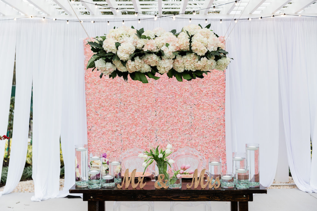 Modern, Whimsical Garden Wedding Decor and Sweetheart Table, Pink Floral Boxwood Wall, White Draping, Ivory, Blush Pink Hanging Floral Arrangement with Greenery, Clear Floating Candles, Gold Mr. and Mrs. Tabletop, White Lilly Bouquet, Sweetheart Table with Ghost Acrylic Chairs, Outdoor Dance floor | Tampa Bay Wedding Planner Special Moments Event Planning | Tampa Bay Wedding Florist Gabro Event Services | Tampa Bay Wedding Rentals A Chair Affair