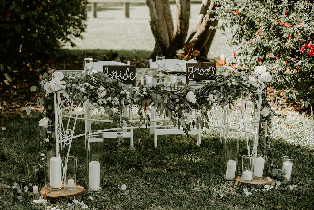 Classic, Organic, Natural Outdoor Garden Wedding Reception Decor, White Table with Greenery Garland and White Flowers, Wooden Bride and Groom Signs, Wood Round Trays with Tall Glass Hurricane Candle Holders and White Candles | Tampa Wedding Venue Davis Islands Garden Club | Tampa Wedding Rentals Elite Events Catering