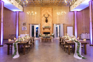 Rustic Country Chic Wedding Indoor Reception Decor, Purple Uplighting Long Wooden Feasting Table, White Table Runner with Greenery Garland, Candlesticks and White Floral Centerpieces, Deer Head Mounted on Wall, White Sweetheart Table | Tampa Bay Wedding Planner Parties A'La Carte | Tampa Wedding Florist Bruce Wayne Florals | Dade City Wedding Venue Covington Farms