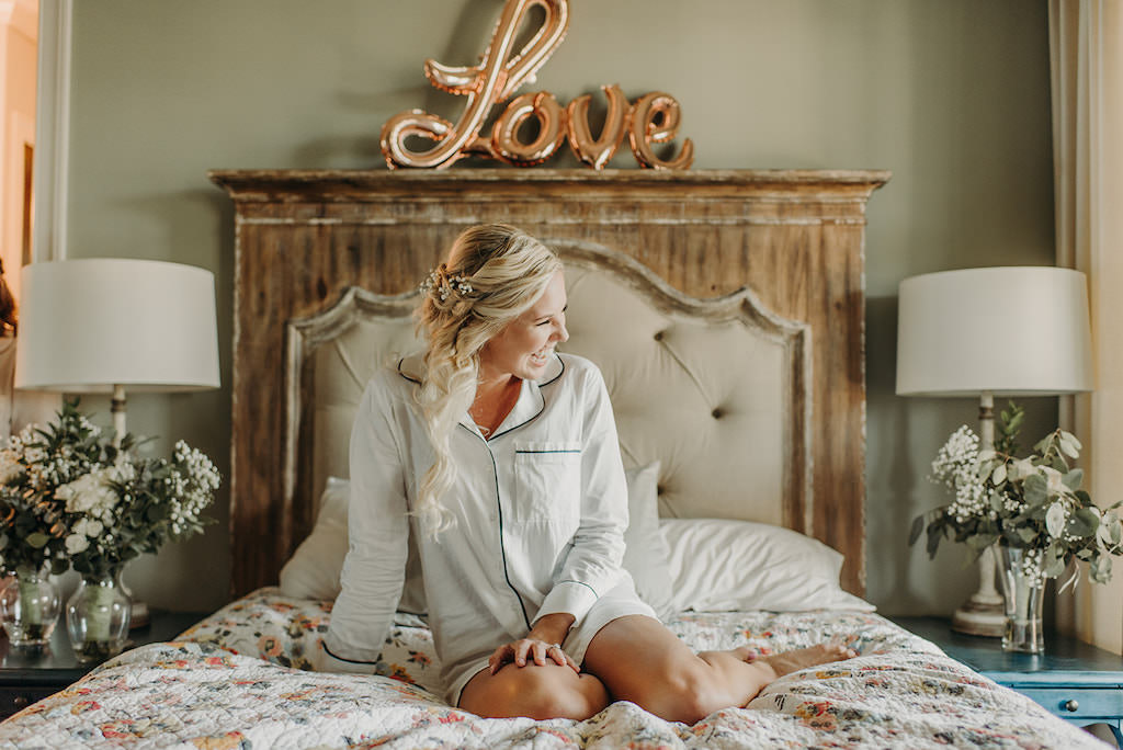 Florida Bride Getting Ready Wedding Portrait in Hotel Suite in White Long Sleeve Pajama, Half Curl Updo with Floral Hair Accessory and Rose Gold Love Balloon Decor | Tampa Bay Wedding Hair and Makeup Femme Akoi