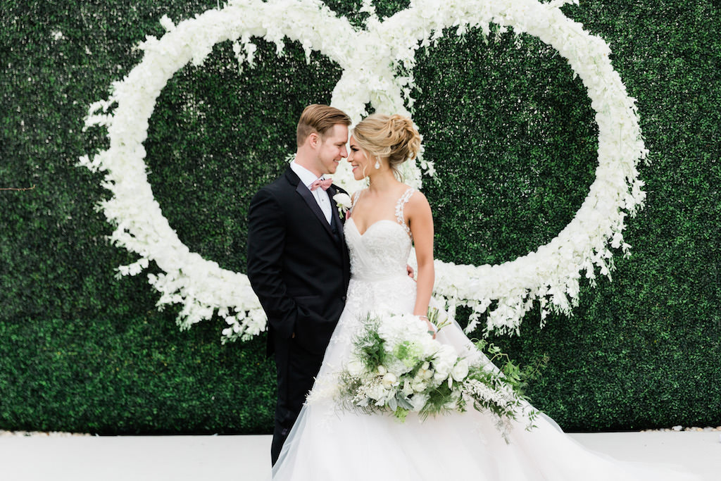 Modern, Elegant Florida Bride and Groom, Wearing White Ballgown Style Wedding Dress with Sweetheart Neckline and Illusion Lace Straps, Carrying Luxurious White and Green Bouquet with Mixed Stems, At Garden Inspired Outdoor Wedding Ceremony, Infinity Ring Hanging Floral Arrangement with White Orchids, Dark Green Boxwood Wall, Wood Cross Back Chairs, Draping | Tampa Bay Wedding Planner Special Moments Event Planning | Tampa Bay Wedding Florist Gabro Event Services | Tampa Bay Hair and Makeup Artists Femme Akoi | Tampa Bay Couture Wedding Dress Boutique Isabel O'Neil Bridal Collection