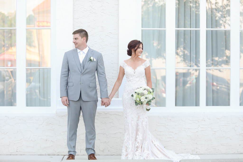 Tampa Bay Bride and Groom Side by Side Holding Hands Wedding Portrait, Groom in Grey Suit with White Tie, Bride in Lace Fitted Off the Shoulder Wedding Dress Holding White, Ivory and Greenery Floral Bouquet | Wedding Photographer Lifelong Photography Studios
