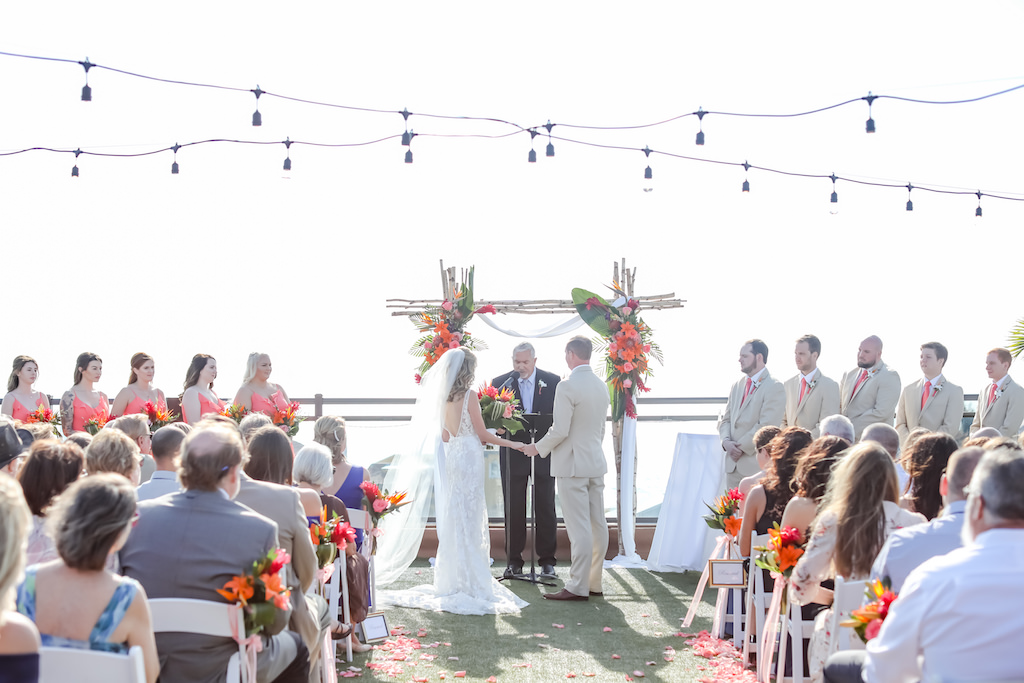Tropical, Beach Inspired Rooftop Waterfront Wedding Ceremony, Gold and Coral Wedding Decor, White Folding Chairs with Bright Flowers in Bouquets, Island Inspired Arch with Vibrant Floral Arrangements, Rooftop String Lighting | Boutique St. Pete Beach Wedding Venue Hotel Zamora | Wedding Rentals By Gabro Event Services | Tampa Bay Wedding Photographer Lifelong Photography Studios