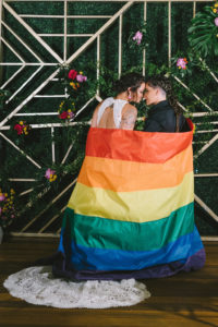 Gay Pride Lesbian Couple Wedding Portrait Wrapped with Rainbow Flag, Modern Geometric Backdrop with Greenery and Red, Yellow and Pink Flower Arrangements | Tampa Bay Wedding Photographer Kera Photography | St. Pete Wedding Planner UNIQUE Weddings & Events