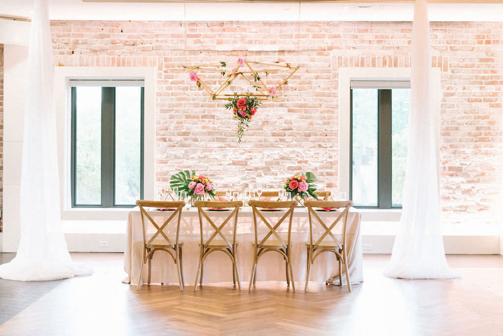 Modern Colorful Tropical Wedding Reception Decor, Feasting Table with Sheer White Linen, Wooden Crossback Chairs, Pink and Yellow Flowers and Green Monstera Palm Leaf Centerpieces, Hanging Gold Geometric Chandelier with Orange, Pink and Greenery Floral Arrangement | Tampa Bay Wedding Photographer Kera Photography | Downtown St. Pete Modern Wedding Venue Red Mesa Events | Wedding Florist and Rentals Gabro Event Services | Wedding Linen and Tabletop Rentals Kate Ryan Event Rentals | Wedding Planner UNIQUE Weddings & Events