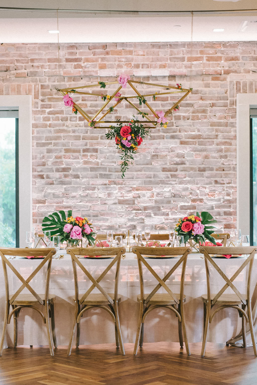 Modern Colorful Tropical Wedding Reception Decor, Feasting Table with Sheer White Linen, Wooden Crossback Chairs, Pink and Yellow Flowers and Green Monstera Palm Leaf Centerpieces, Hanging Gold Geometric Chandelier with Orange, Pink and Greenery Floral Arrangement | Tampa Bay Wedding Photographer Kera Photography | Downtown St. Pete Modern Wedding Venue Red Mesa Events | Wedding Florist and Rentals Gabro Event Services | Wedding Linen and Tabletop Rentals Kate Ryan Event Rentals | Wedding Planner UNIQUE Weddings & Events