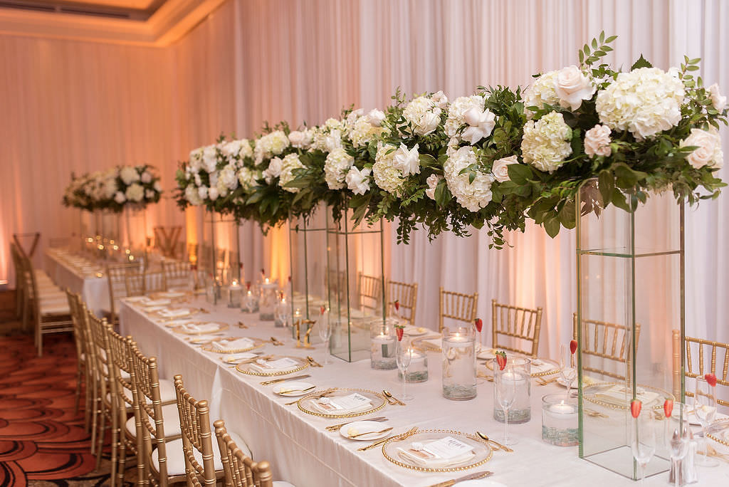 Elegant Classic Romantic Wedding Reception Decor, Long Feasting Table with White Linens, Gold Chiavari Chairs, Clear Glass and Gold Beaded Rimmed Chargers, Tall Glass Rectangular Vase with White Hydrangeas, Ivory and Blush Pink Flowers with Greenery Floral Arrangement | Tampa Bay Wedding Photographer Kristen Marie Photography | Clearwater Beach Wedding Florist, Rentals, Linens, and Lighting Gabro Event Services | Wedding Day of Coordinator Special Moments Event Planning
