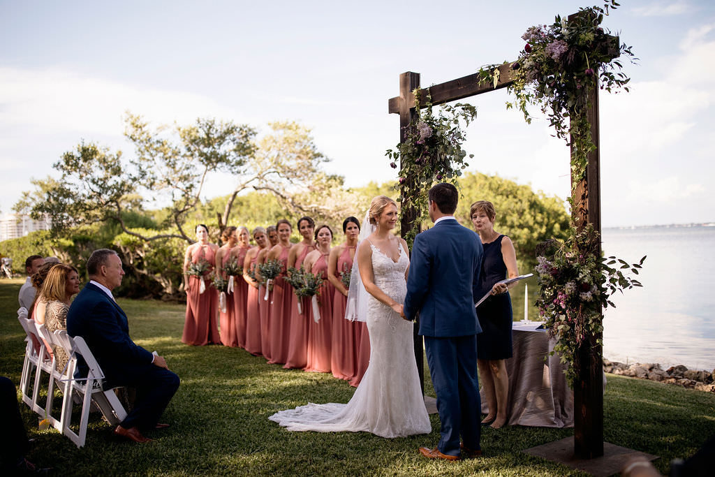 Florida Bride and Groom Exchanging Wedding Vows During Ceremony Under Wooden Arch with Greenery and Purple Floral Arrangements Waterfront Outdoor Wedding Portrait | Wedding Venue The Resort at Longboat Key Club