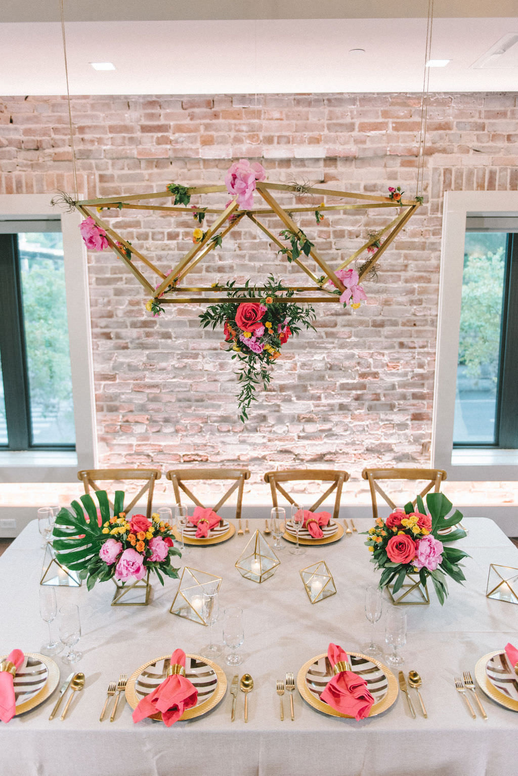Modern Colorful Tropical Wedding Reception Decor, Feasting Table with Sheer White Linen, Wooden Crossback Chairs, Pink and Yellow Flowers and Green Monstera Palm Leaf Centerpieces, Hanging Gold Geometric Chandelier with Orange, Pink and Greenery Floral Arrangement, Gold Chargers and Plates with Bright Pink Linen with Gold Napkin Ring | Tampa Bay Wedding Photographer Kera Photography | Downtown St. Pete Modern Wedding Venue Red Mesa Events | Wedding Florist and Rentals Gabro Event Services | Wedding Linen and Tabletop Rentals Kate Ryan Event Rentals | Wedding Planner UNIQUE Weddings & Events