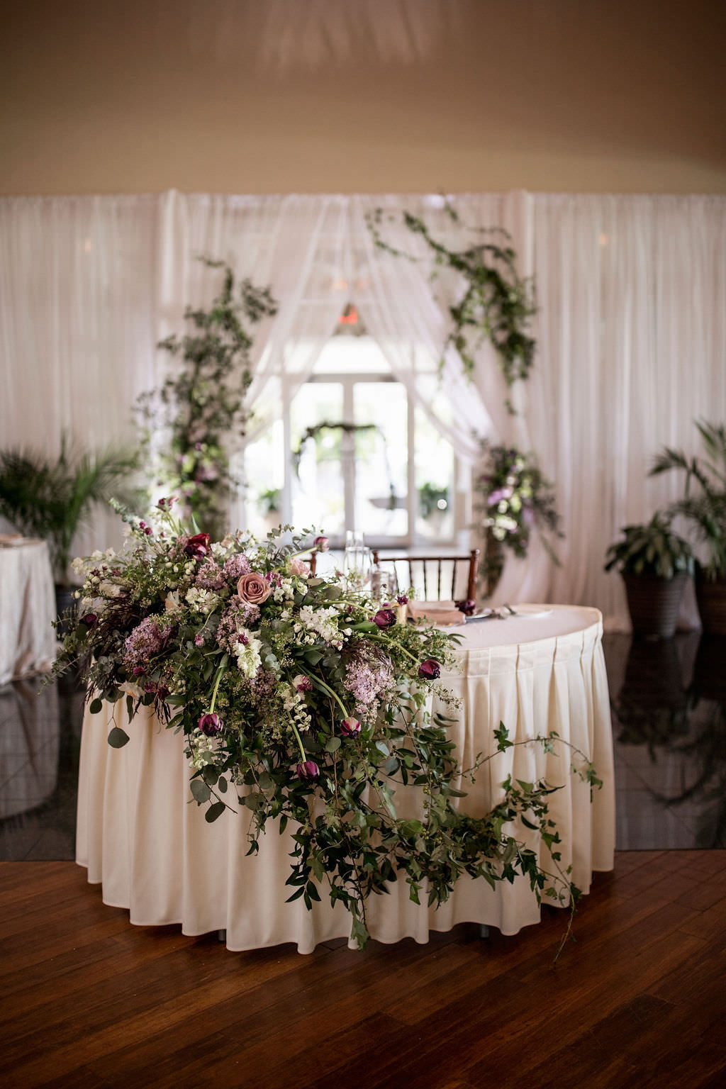 Elegant Classic Wedding Reception Decor, Sweetheart Table with White Linen, Greenery Garland and Lilac, Purple, Plum, and Ivory Floral Centerpiece and White Linen Backdrop
