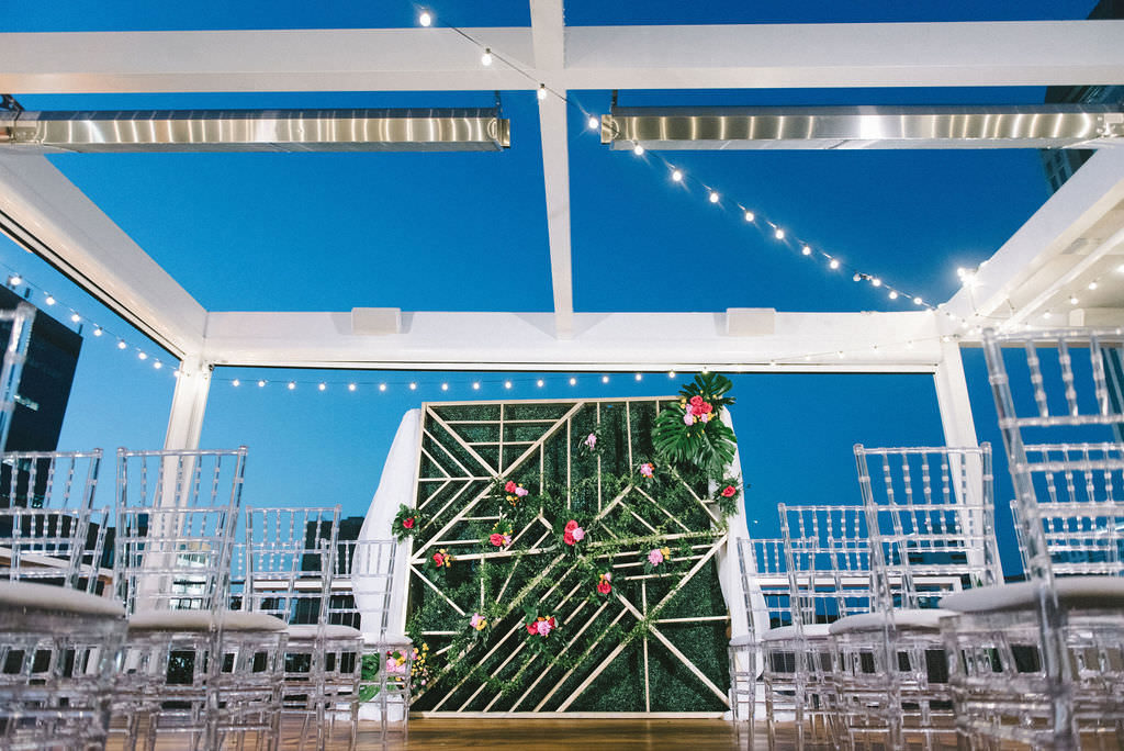 Modern Rooftop Wedding Ceremony Decor, Hanging String Lights, Geometric and Greenery Wall Backdrop with Colorful Red and Pink Flowers, Ghost Clear Acrylic Chiavari Chairs | Tampa Bay Wedding Photographer Kera Photography | Downtown Rooftop St. Pete Wedding Venue Red Mesa Events | Wedding Planner UNIQUE Weddings & Events | Wedding Florist and Rentals Gabro Event Services