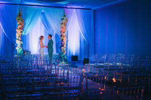 Florida Bride and Groom Wedding Ceremony Portrait, Blue Uplighting, White Draping Ceremony Backdrop, Tall Large Colorful Hydrangeas Floral Pillars, Ghost Acrylic Clear Chiavari Chairs Decor | Tampa Bay Boutique Hotel Wedding Venue Hotel Alba | Wedding Planner Special Moments Event Planning | Wedding Rentals and Chair Decor Gabro Event Services | Styled Shoot