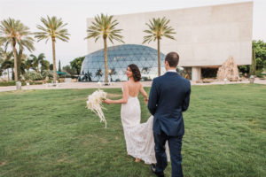 Tampa Bride and Groom Wedding Portrait, Groom Holding Train of Bride's Hayley Paige Lace Open Back Wedding Dress with St. Petersburg Salvador Dali Museum Glass Dome Wedding Venue Backdrop