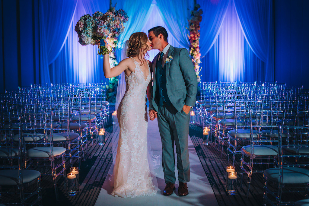 Intimate Bride and Groom Wedding Ceremony Exit Recessional Wedding Portrait, Bride in Lace and Illusion Deep V Neckline Fitted Silhouette Wedding Dress Holding Colorful Hydrangeas Floral Bridal Bouquet, Groom in Grey Suit, Clear Ghost Acrylic Chiavari Chairs, Blue Uplighting and Linen Drapery Backdrop | Tampa Bay Boutique Hotel Wedding Venue Hotel Alba | Wedding Attire Truly Forever Bridal | Wedding Hair and Makeup Michele Renee the Studio | Wedding Planner Special Moments Event Planning | Wedding Rentals and Chair Decor Gabro Event Services | Styled Shoot