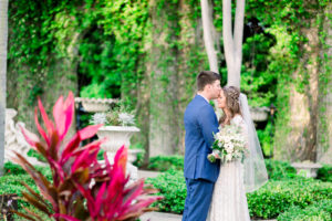 Boho Inspired Florida Bride and Groom in Outdoor Garden Courtyard, Groom in Navy Suit, Bride Holding White Floral Bouquet with Greenery Wedding Portrait | Tampa Bay Wedding Photographers Shauna and Jordon Photography