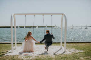 Fun Creative Tampa Bay Bride and Groom Holding Hands on Swing , St. Pete Waterfront Wedding Portrait | UNIQUE Weddings + Events