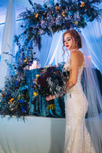 Modern Contemporary Moody and Dramatic Wedding Styled Shoot Bridal Portrait, Sweetheart Table with Blue Linen, Unique Purple, Blue, Orange and Greenery Floral Arrangements, Blue Uplighting and Linen Drapery, Bride in Lace and Illusion Fitted Silhouette Wedding Dress Holding Dark Purple Floral Bouquet | Tampa Bay Wedding Planner Special Moments Event Planner | Wedding Attire Truly Forever Bridal | Wedding Hair and Makeup Michele Renee the Studio | Styled Shoot