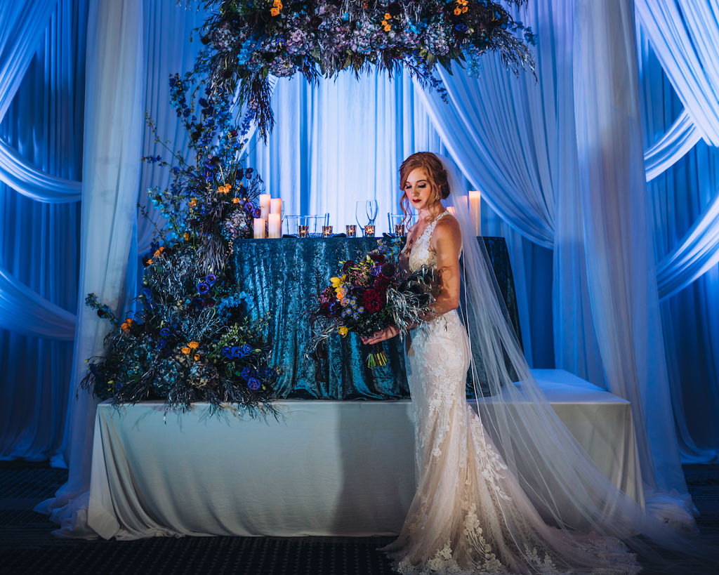 Modern Contemporary Moody and Dramatic Wedding Styled Shoot Bridal Portrait, Sweetheart Table with Blue Linen, Unique Purple, Blue, Orange and Greenery Floral Arrangements, Blue Uplighting and Linen Drapery, Bride in Lace and Illusion Fitted Silhouette Wedding Dress Holding Dark Purple Floral Bouquet | Tampa Bay Wedding Planner Special Moments Event Planner | Wedding Attire Truly Forever Bridal | Wedding Hair and Makeup Michele Renee the Studio | Styled Shoot