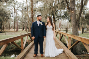 Rustic Chic Bride and Groom on Wooden Bridge Outdoor Wedding Portrait, Bride in Lace and Illusion V Neckline Wedding Dress, Groom in Blue Suit with Blush Pink Tie | Parrish Wedding Venue Rafter J Ranch